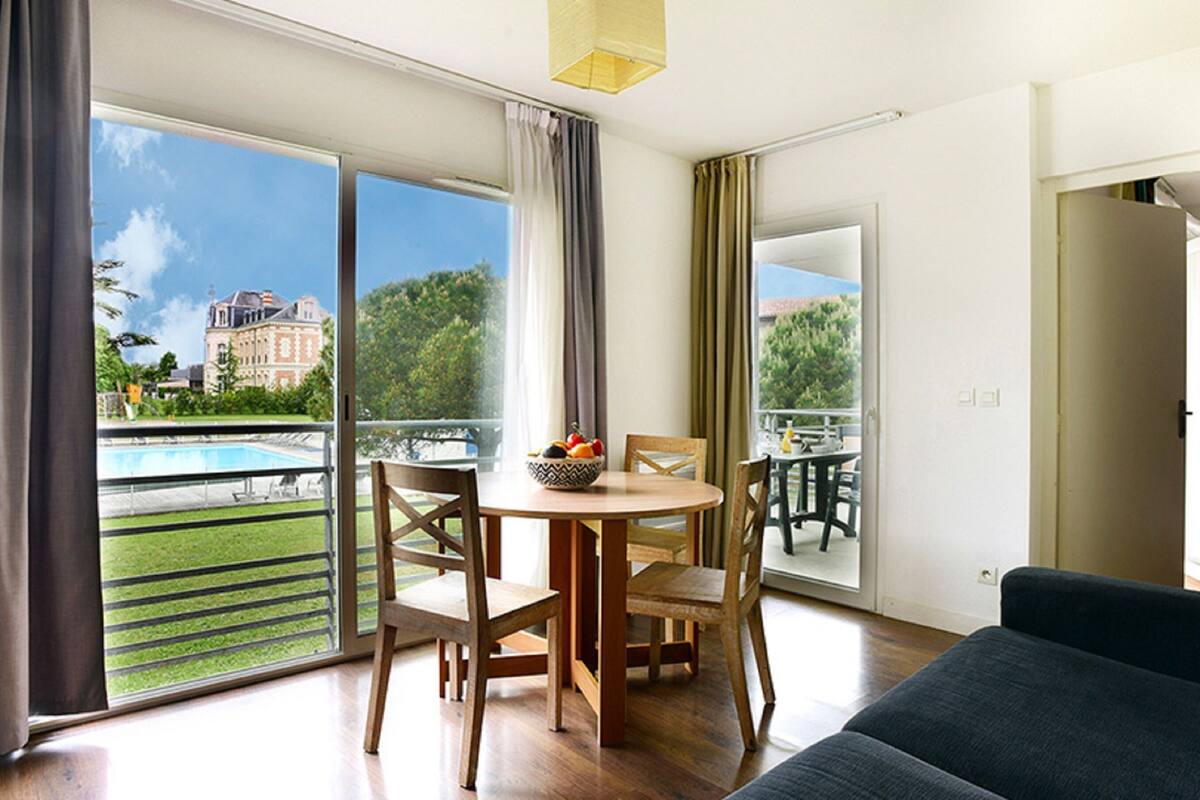 Un salon moderne avec un coin repas, de grandes portes-fenêtres coulissantes donnant sur une piscine et un jardin, et une vue sur un bâtiment historique.