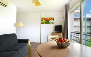 Una habitación de hotel moderna con un sofá, una mesa de comedor con frutero, un televisor y un cuadro en la pared.