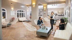 A spacious lounge area with white chairs, a coffee table, and a bar area in the background. - Baia D'Oro Hotel (Licata)