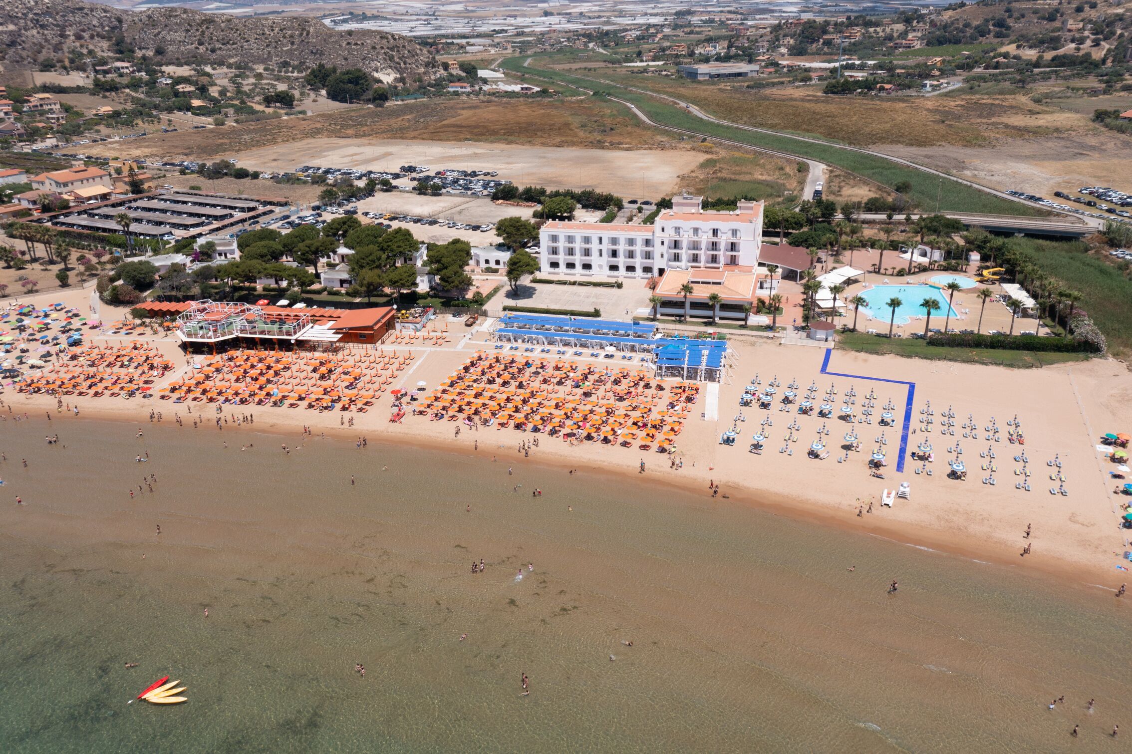 An aerial view of a beach resort with multiple sun loungers and umbrellas, a swimming pool, and a large building with a red roof.