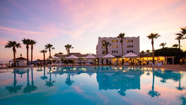 A large outdoor swimming pool with lounge chairs and umbrellas, surrounded by palm trees and a building with multiple windows. - Baia D'Oro Hotel (Licata)