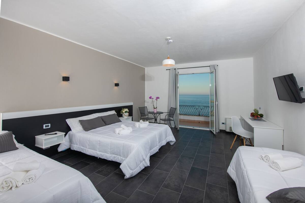 A hotel room with two beds, a desk, a chair, a TV, and a balcony with a view of the sea.