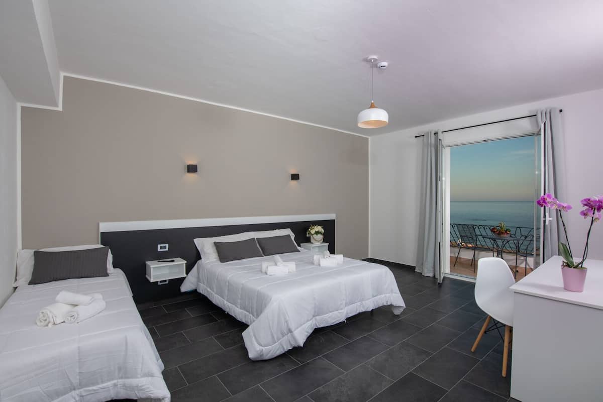 A hotel room with two beds, a desk, and a balcony with a view of the sea.