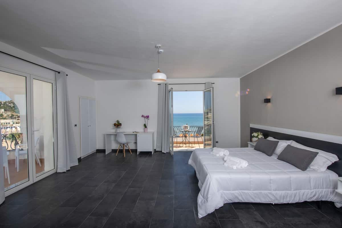 A modern bedroom with a large bed, a desk, and a view of the sea.