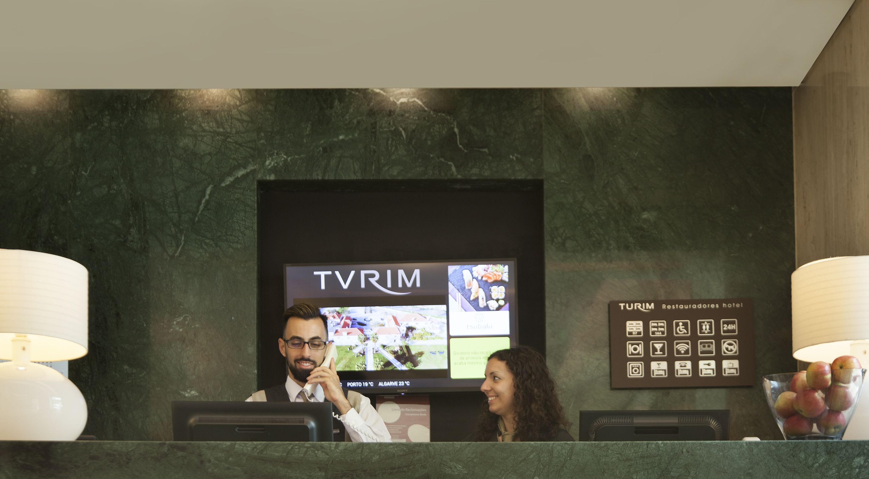 Una reception d'albergo con uno schermo televisivo che mostra 'TVRIM' e un cartello che indica 'Turim Restaurante hotel'.