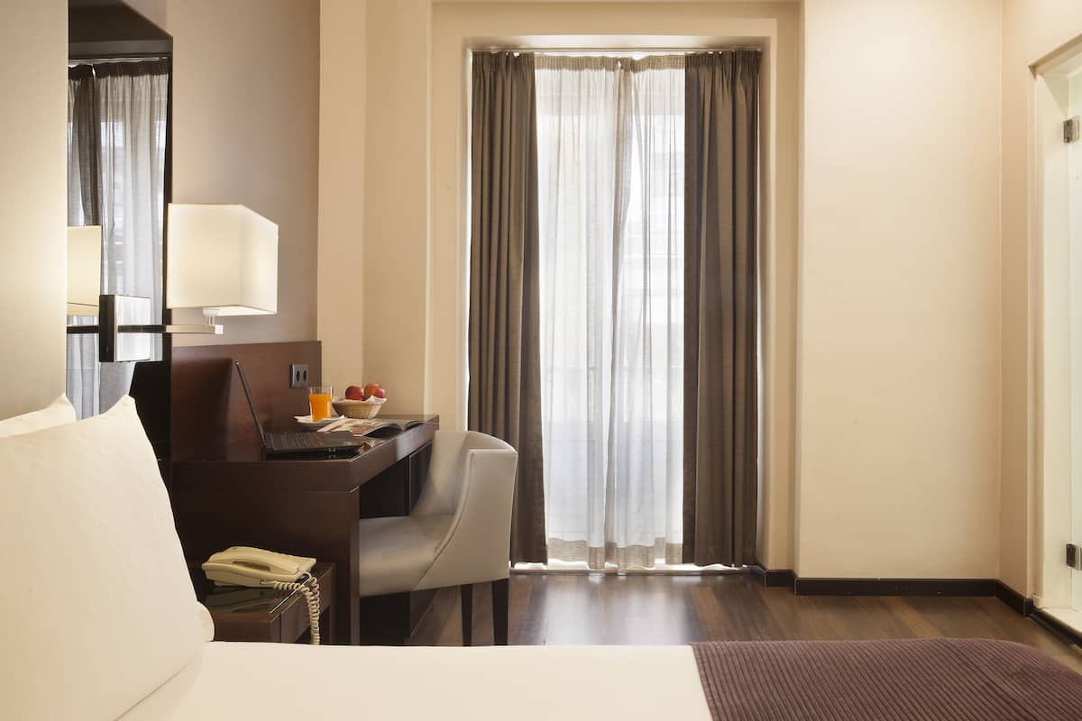 Hotelkamer met een bed, een bureau met lamp, een stoel, een telefoon en een raam met gordijnen.