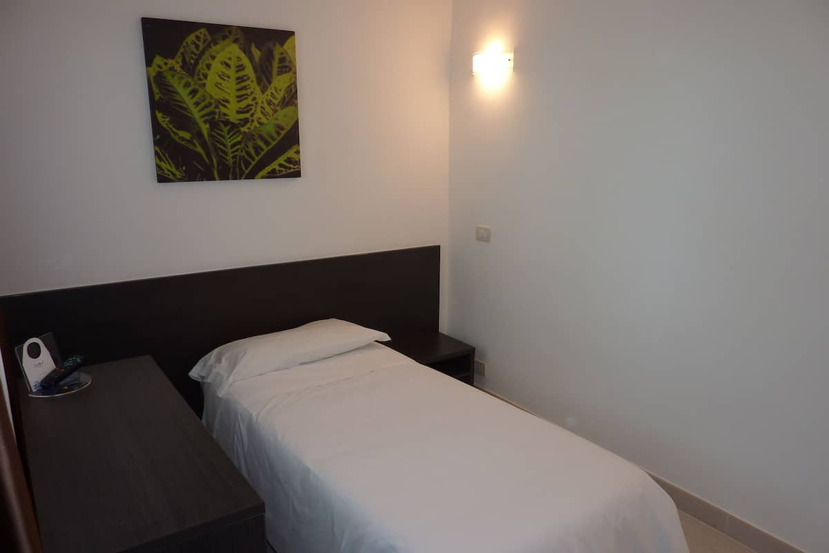 Ein kleines, ordentlich bezogenes Bett mit weißer Bettdecke in einem schlichten Zimmer, versehen mit einem dunklen Kopfteil.