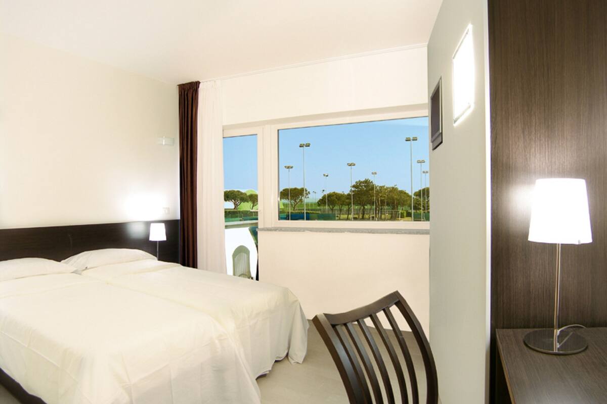 Ein Hotelzimmer mit Bett, Schreibtisch, Stuhl, Fenster mit Blick auf Bäume und einen Tennisplatz, sowie einer Lampe.