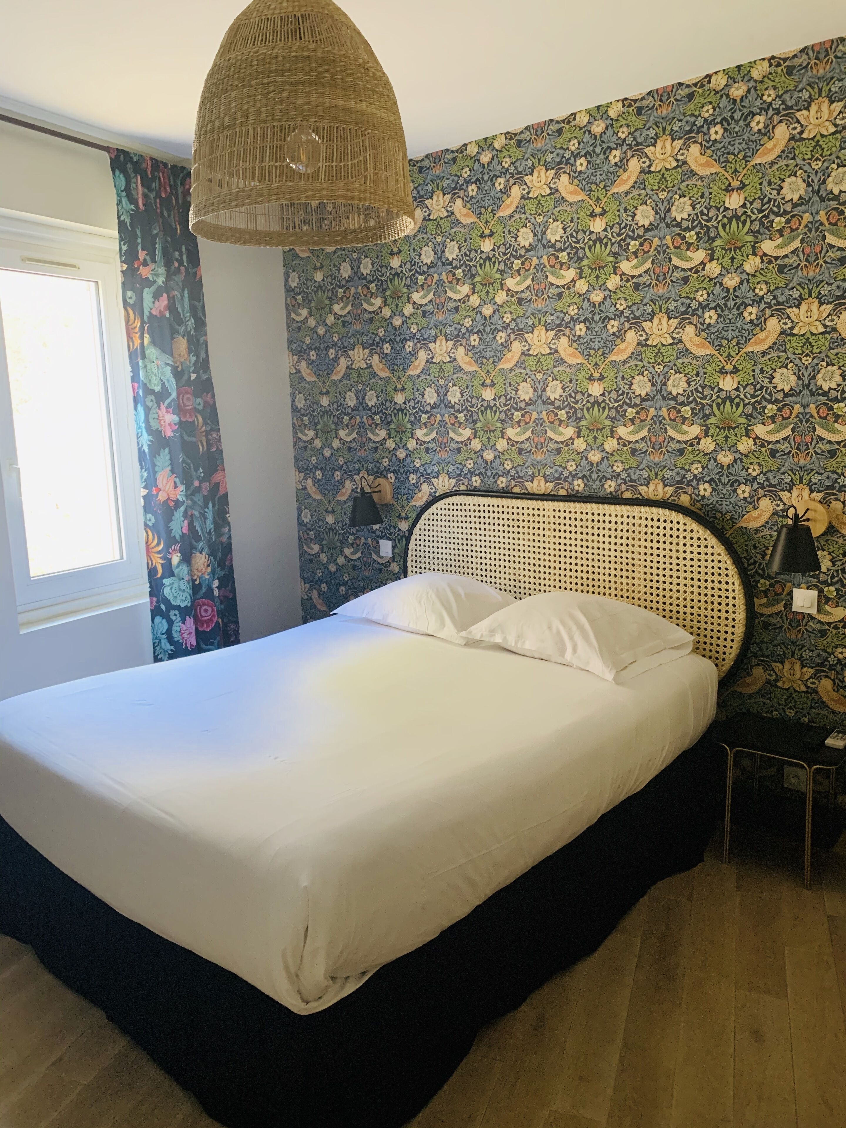 A bed with a woven headboard, a window with floral curtains, and a wall with a bird-patterned wallpaper.