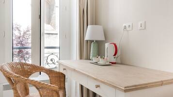 A room with a wicker chair, a white desk with a lamp, and a red kettle.