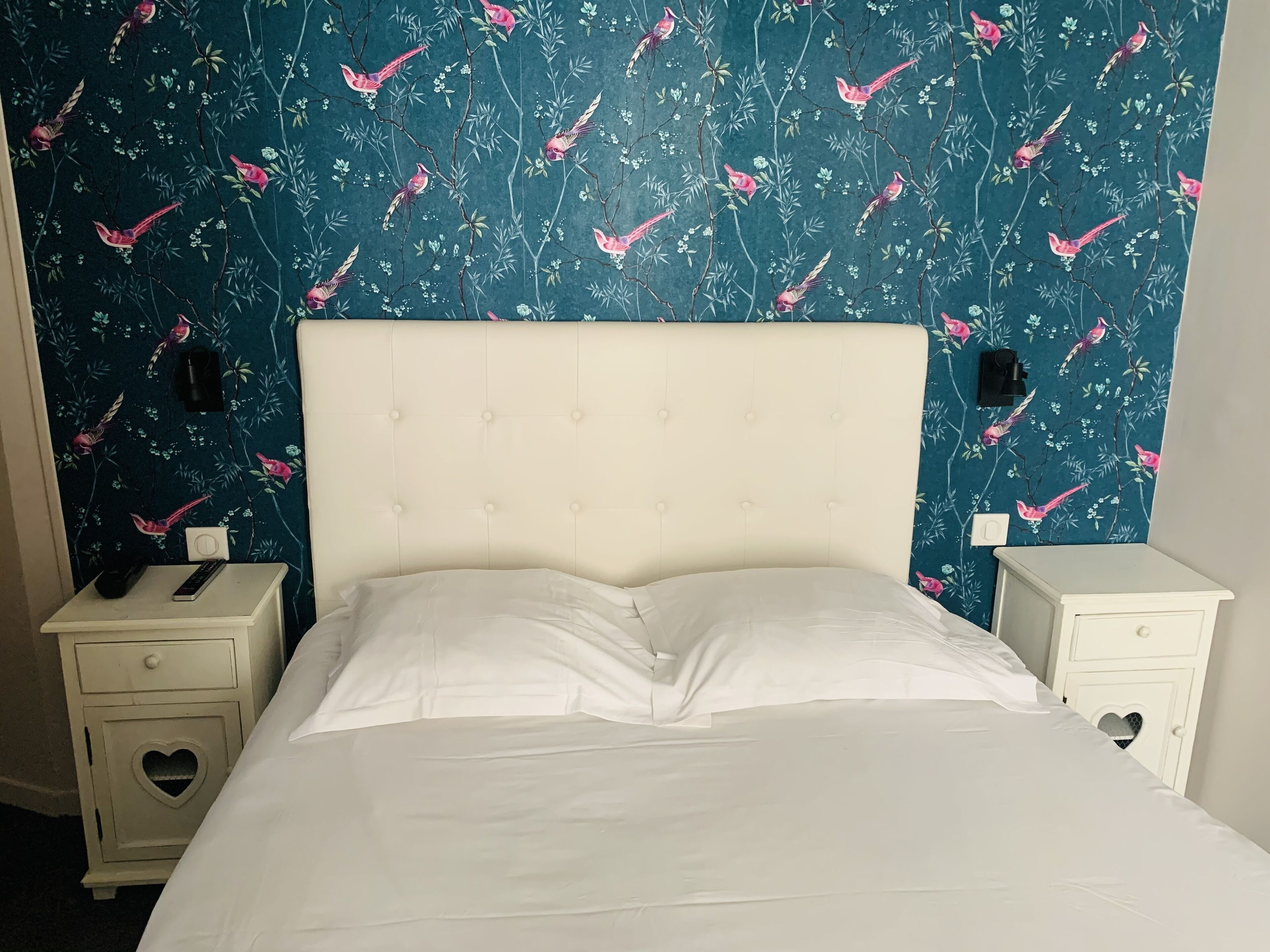 Un lit avec du linge de lit blanc, une tête de lit capitonnée et un papier peint à motifs floraux avec des oiseaux. Il y a deux tables de chevet avec des découpes en forme de cœur, et l’une d’entre elles porte une télécommande.