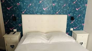 A bed with white linens, a tufted headboard, and a floral wallpaper with birds. There are two bedside tables with heart-shaped cutouts, one with a remote control on it.