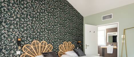A hotel room with a large bed, a wooden headboard, a bedside table, a crib, a bathroom with a mirror, and a green leafy wallpaper.