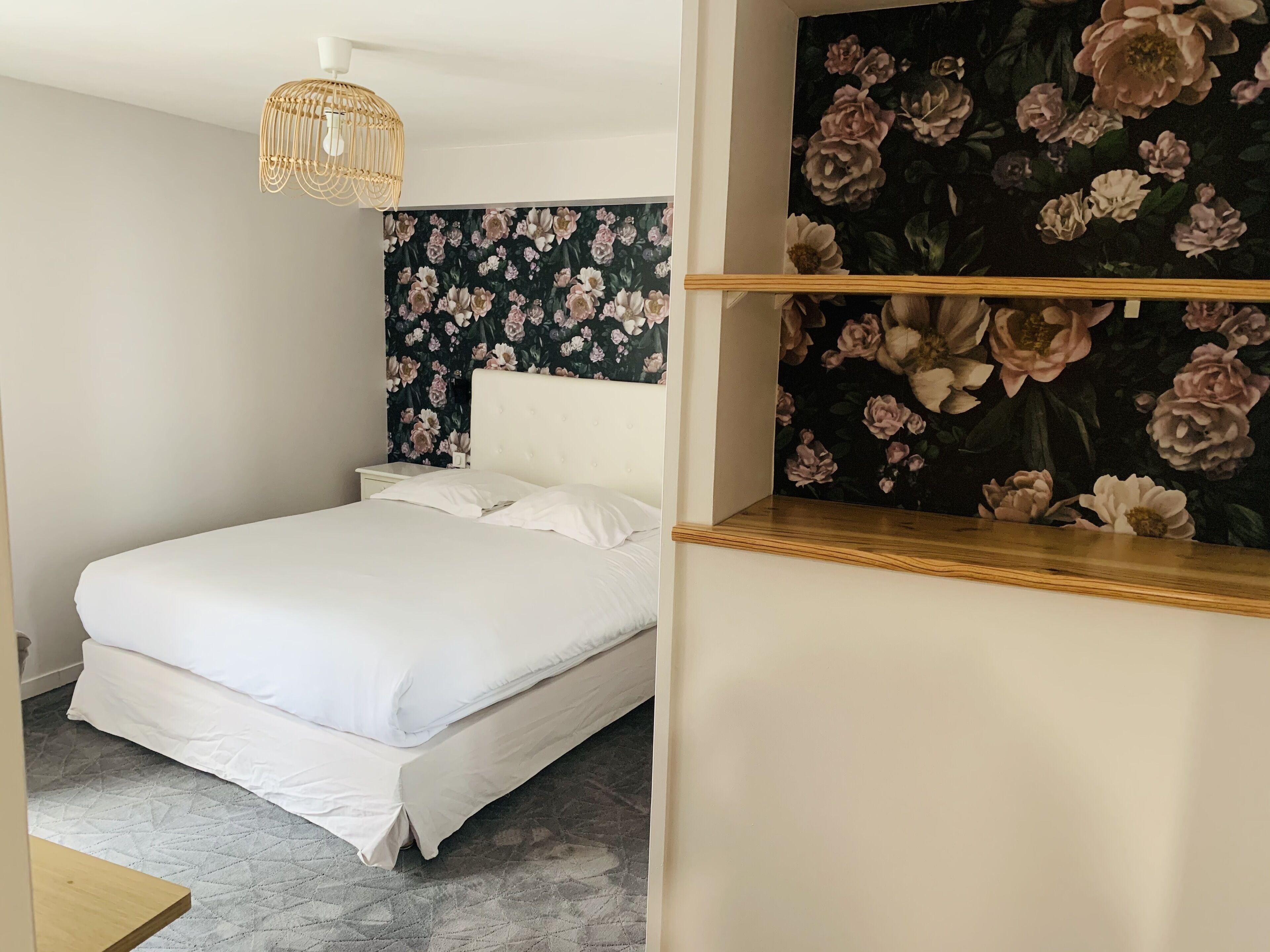 Un lit bien fait, avec du linge de lit blanc, dans une pièce dont le papier peint est à motifs floraux et qui est agrémenté d’une tête de lit en bois.