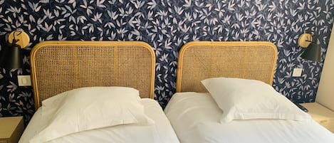 A room with two beds, each with white bedding and a woven headboard. The walls have a dark blue floral wallpaper.