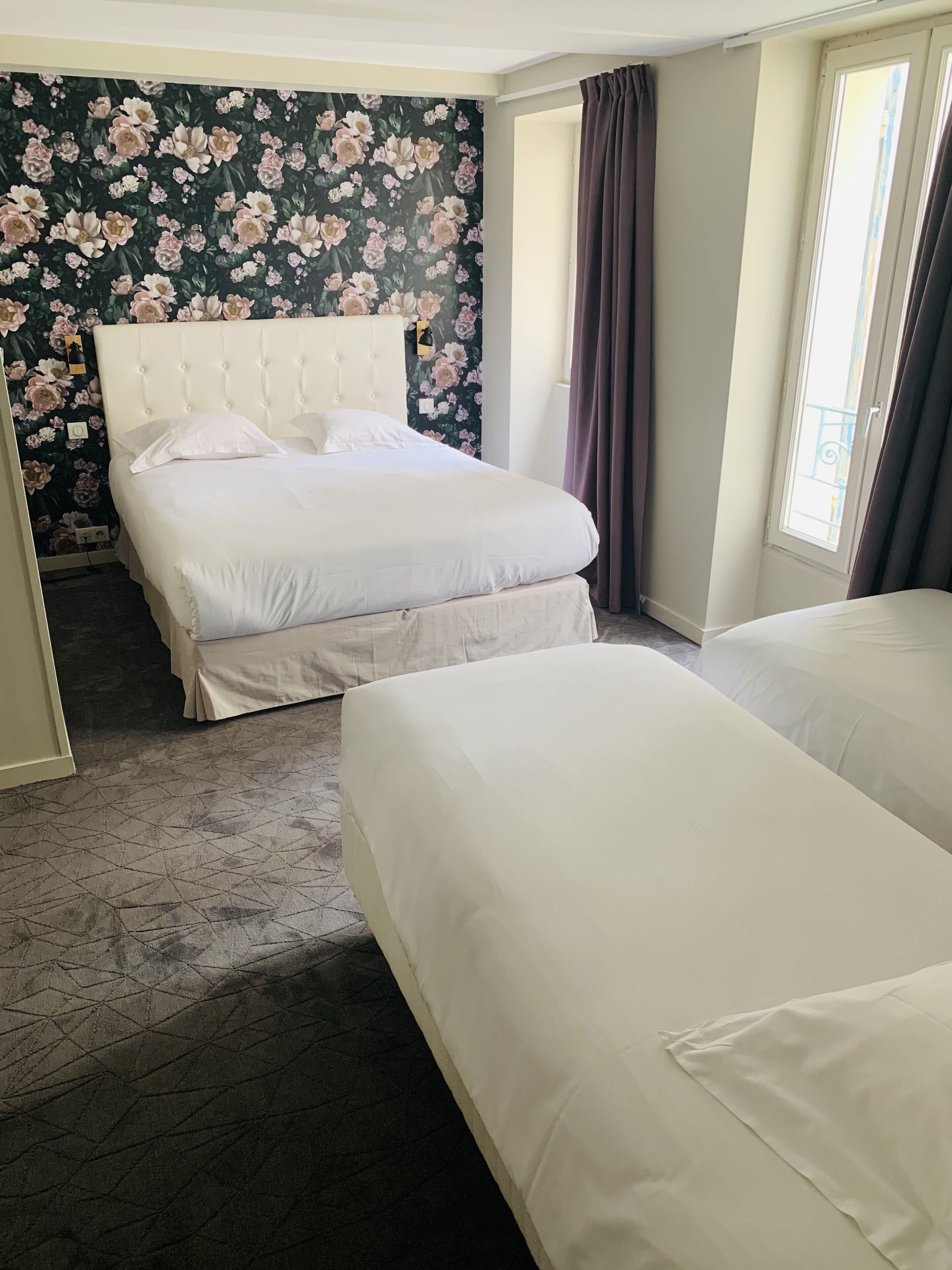 A hotel room with a large bed, a smaller bed, a floral wallpaper, and a window with curtains.