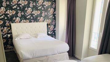 A hotel room with a large bed, a smaller bed, a floral wallpaper, and a window with curtains.