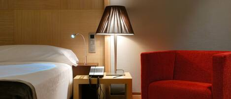 A hotel room with a bed, a desk with a lamp, a telephone, and a red chair.