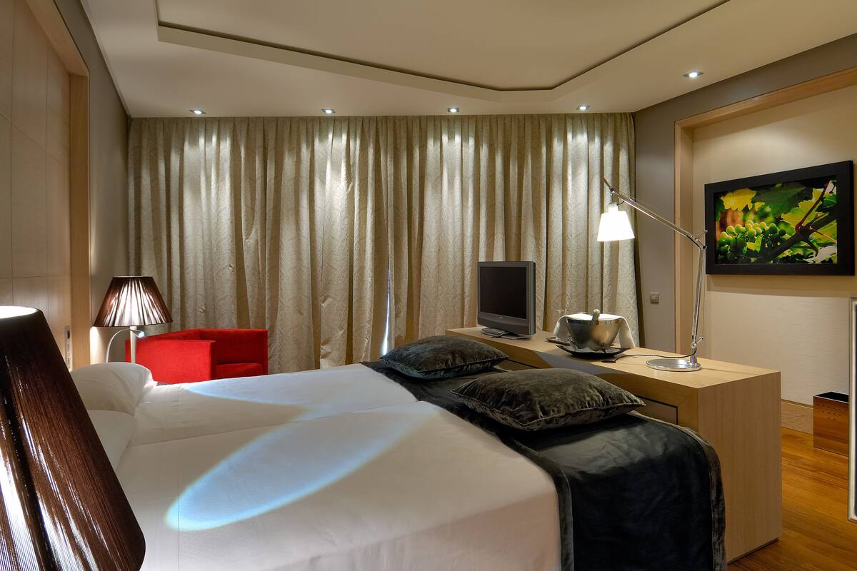 Una habitación de hotel moderna con una cama grande, un escritorio con computadora, una silla roja, una lámpara y un cuadro en la pared.