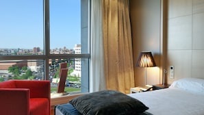 Une chambre d’hôtel avec une grande fenêtre, un lit, une table de chevet, une lampe et une chaise rouge.