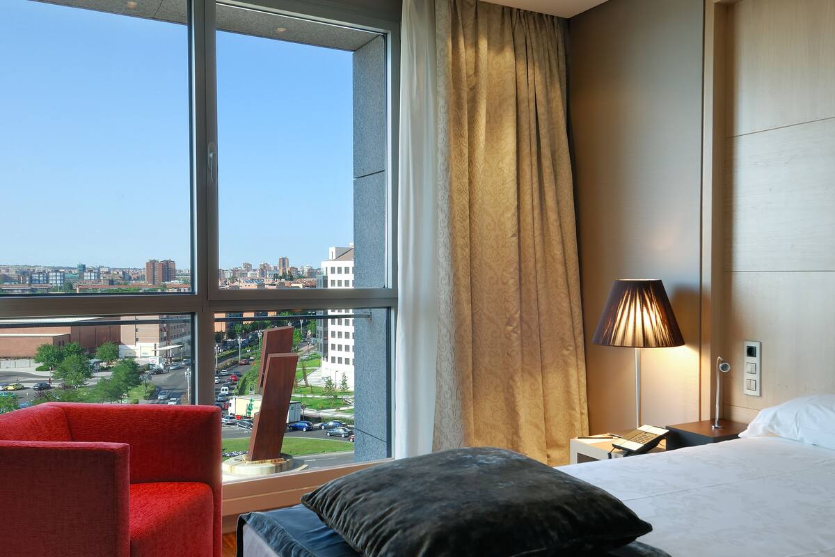 Una habitación de hotel con un ventanal grande, una cama, una mesita de noche, una lámpara y una silla roja.