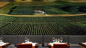 Un vignoble avec des rangées de vignes et un bâtiment au centre. Au premier plan, on aperçoit des tables avec des verres à vin et des assiettes.