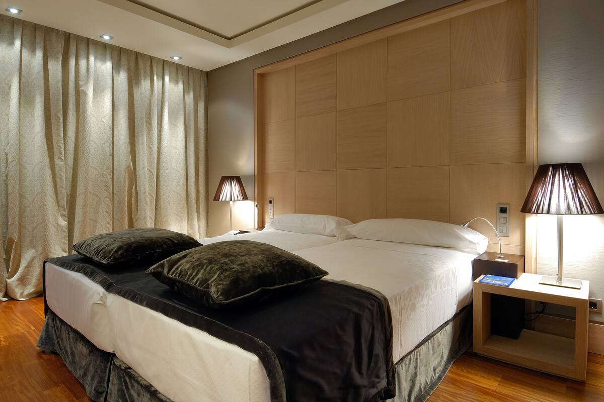 Una cama doble con dos almohadas oscuras, un cabecero de madera, lámparas de noche y una mesita de noche con un libro.