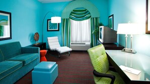 Une chambre d’hôtel moderne aux couleurs turquoise, avec un canapé, un fauteuil, un bureau, une lampe et un téléviseur.