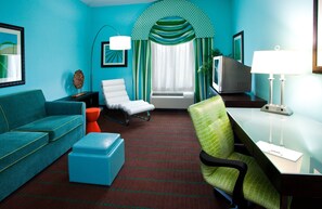 A modern hotel room with a teal color scheme, a sofa, a chair, a desk, a lamp, and a television. - Holiday Inn & Suites Ocala Conference Center by IHG (Ocala)