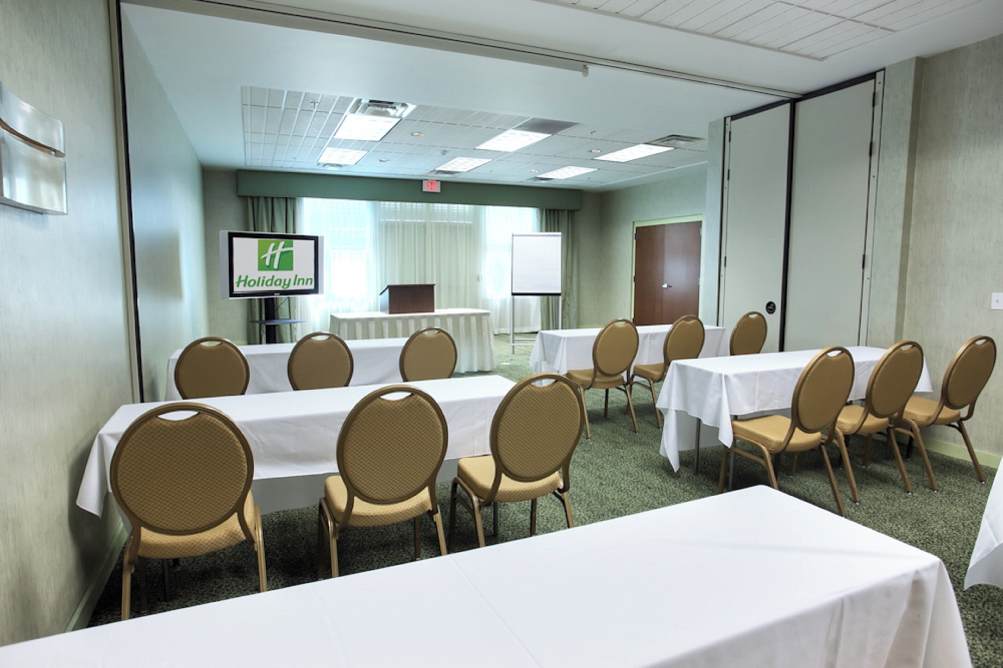 A conference room with multiple tables and chairs, a screen displaying the Holiday Inn logo, and a whiteboard in the background.