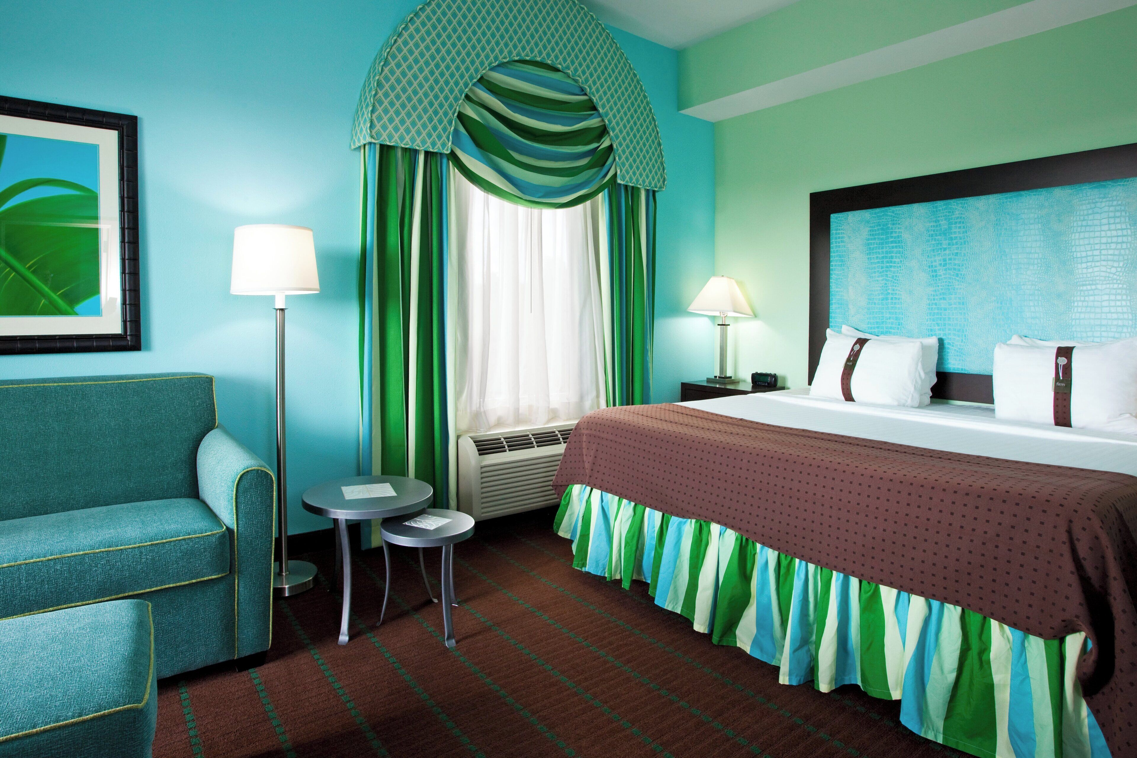 Una habitación de hotel con una cama, una silla, una mesita, una lámpara y un cuadro en la pared.