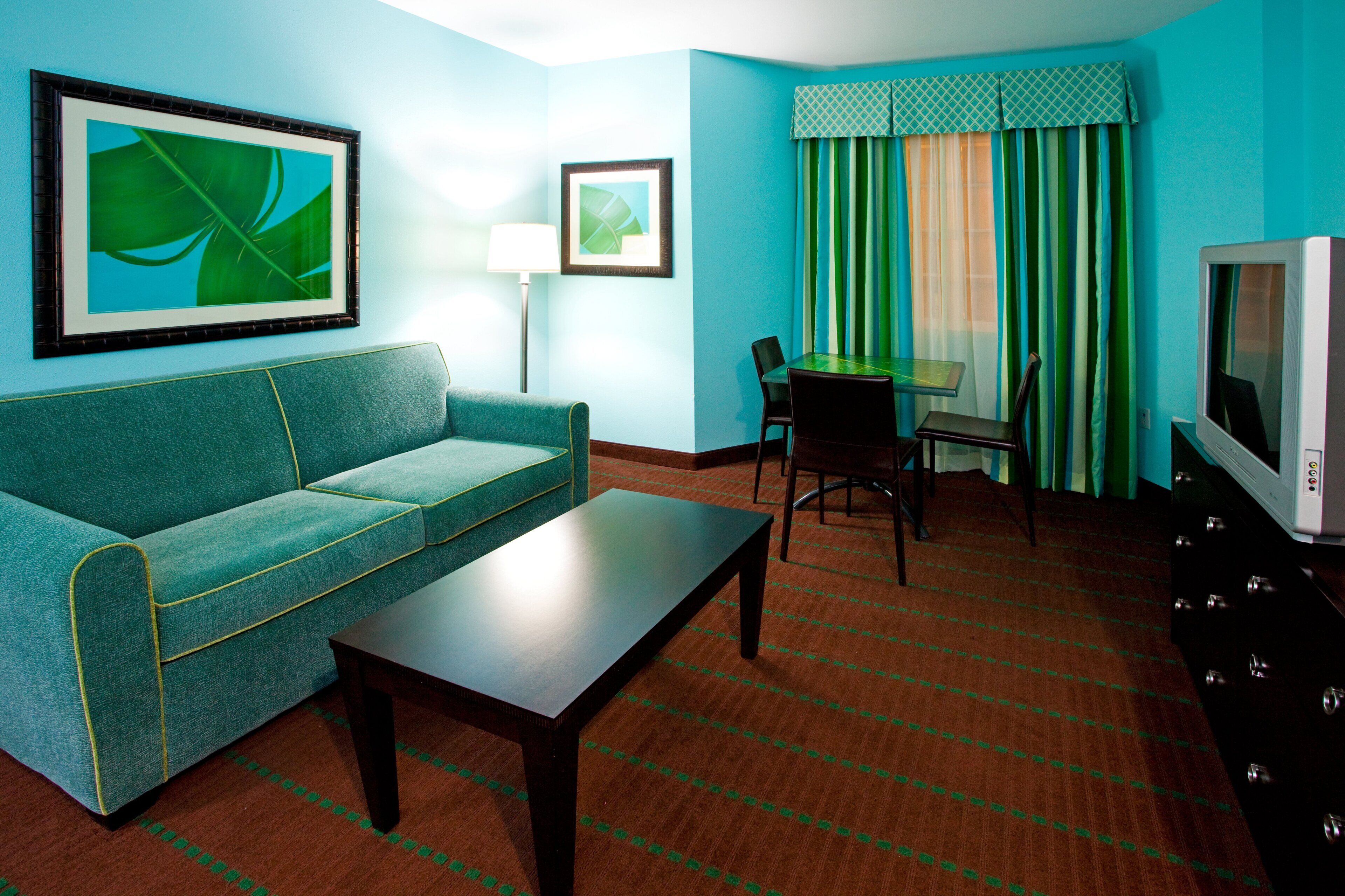 Une chambre d’hôtel avec un canapé de couleur turquoise, une petite table, un téléviseur, un bureau avec une chaise et une fenêtre avec des rideaux à rayures.