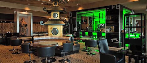 A spacious lounge area with a large central seating arrangement, a decorative wall with a clock, and a green-lit bar area.