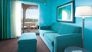 Une pièce avec un canapé de couleur turquoise, un pouf assorti, une petite table, un tableau encadré au mur et une vue sur le balcon par la porte.