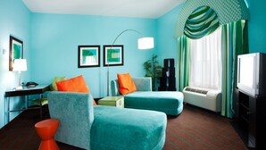Una stanza dalle pareti color acquamarina, con una poltrona color acquamarina, uno sgabello arancione, una scrivania con una lampada, una televisione e un'ampia finestra con tende.