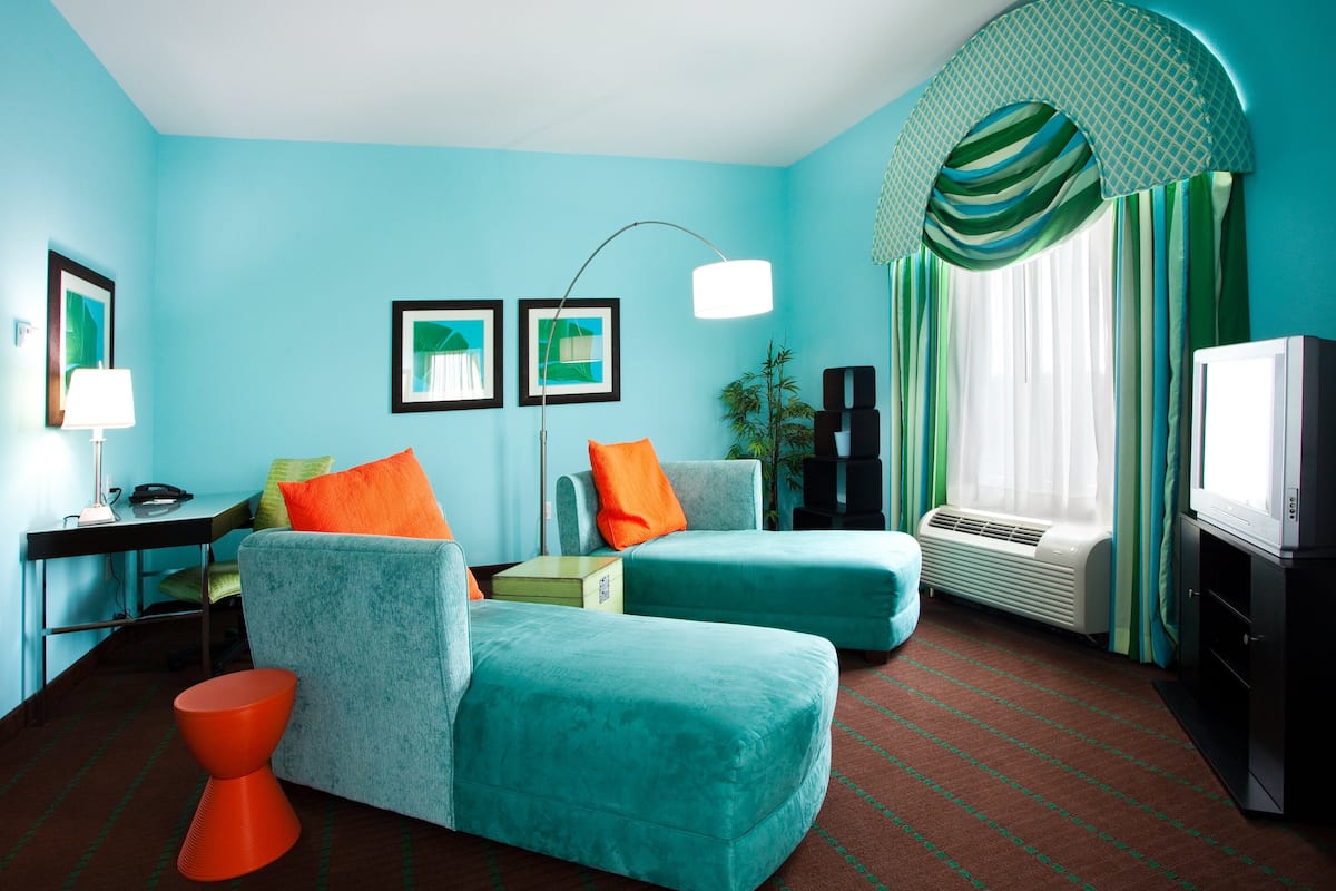Ein Zimmer mit türkisfarbenen Wänden, einer türkisfarbenen Sofagarnitur, einem orangefarbenen Hocker, einem Schreibtisch mit Lampe, einem Fernseher und einem großen Fenster mit Vorhängen.