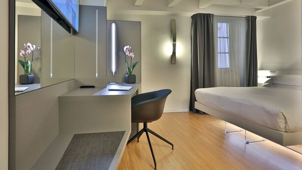 A modern hotel room with a bed, a desk, a chair, a TV, and two potted orchids. - Urban Hotel Design (Trieste)