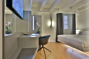 A modern hotel room with a bed, a desk, a chair, a TV, and two potted orchids.