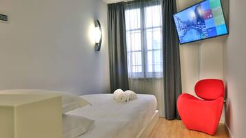 A hotel room with a bed, a red chair, a TV on the wall, and a window with curtains.