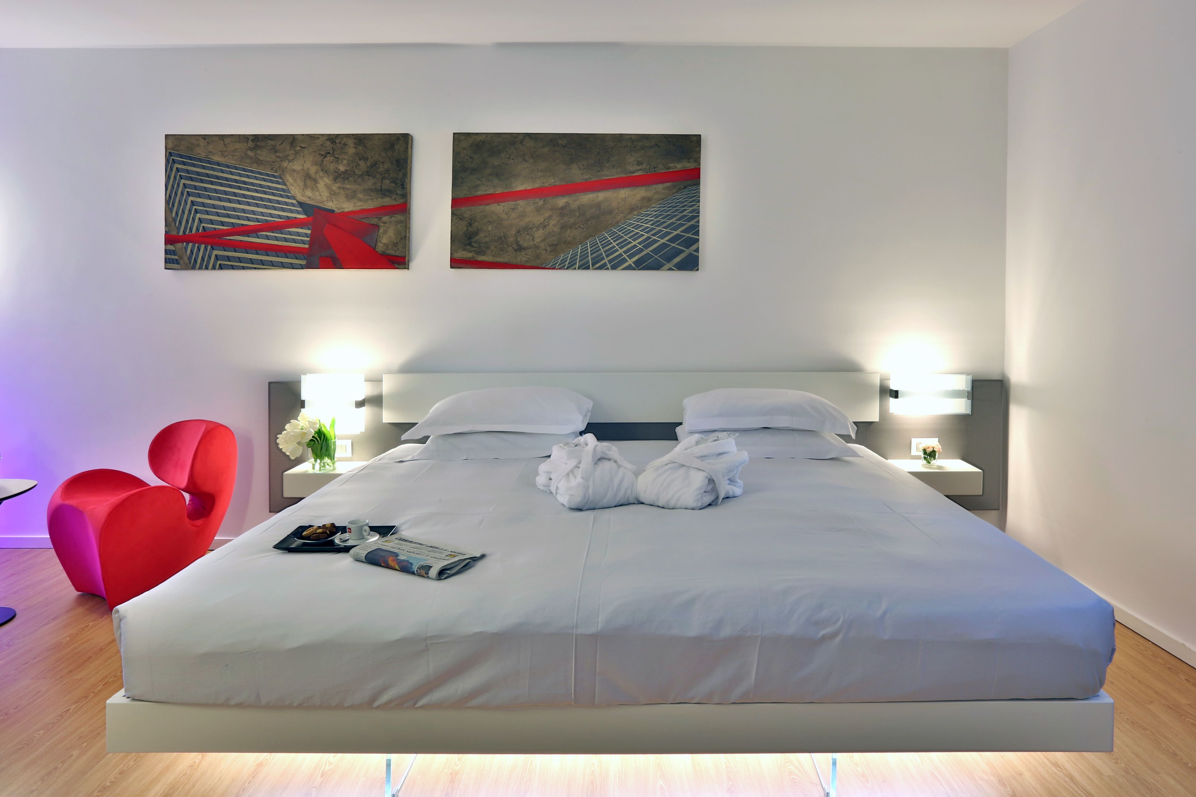 A modern hotel room with a large bed, two wall-mounted artworks, a red chair, and a nightstand with a lamp.