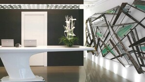 A modern reception area with a white reception desk, a potted plant, and a decorative wall piece. - Urban Hotel Design (Trieste)