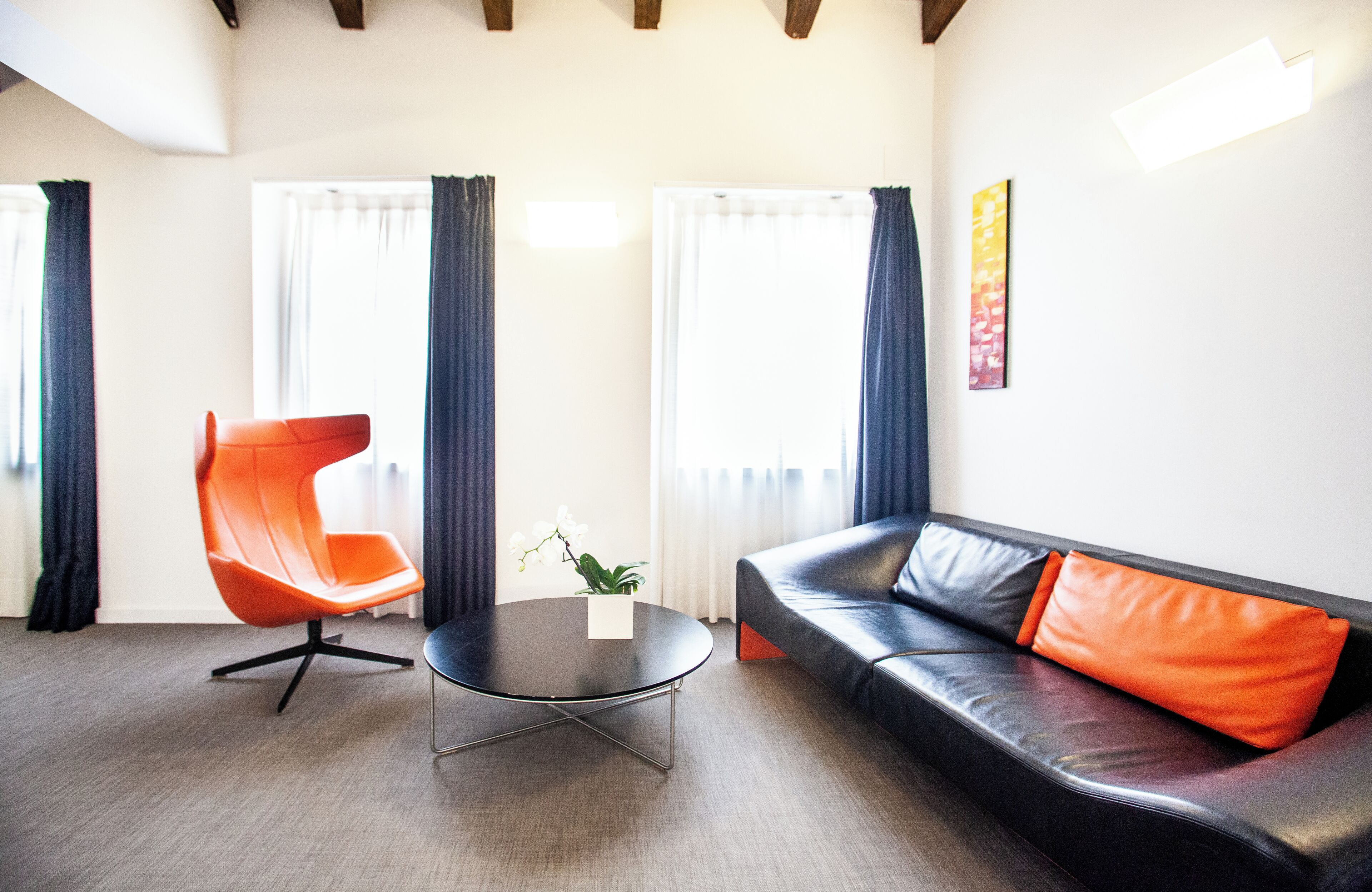 A modern living room with a leather sofa, an orange chair, a round coffee table, and large windows with curtains.