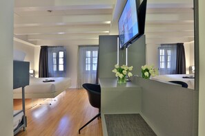A hotel room with a bed, a desk with a computer, a chair, a TV mounted on the wall, and a window with curtains.