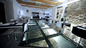 A modern dining area with glass tables and chairs, a kitchen in the background, and a stone wall on one side. - Urban Hotel Design (Trieste)