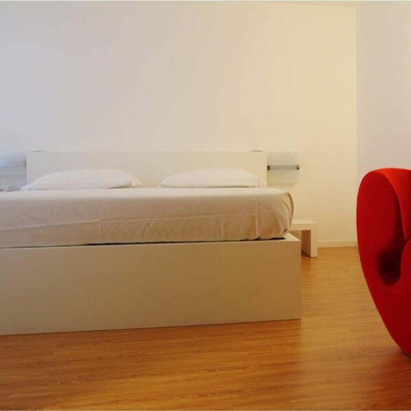 A minimalist bedroom with a white bed, a red chair, and a wooden floor.
