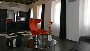 Una sala de estar moderna con un sillón rojo, una mesa de centro redonda y un sofá. Se observa un televisor de pantalla plana y un comedor al fondo.