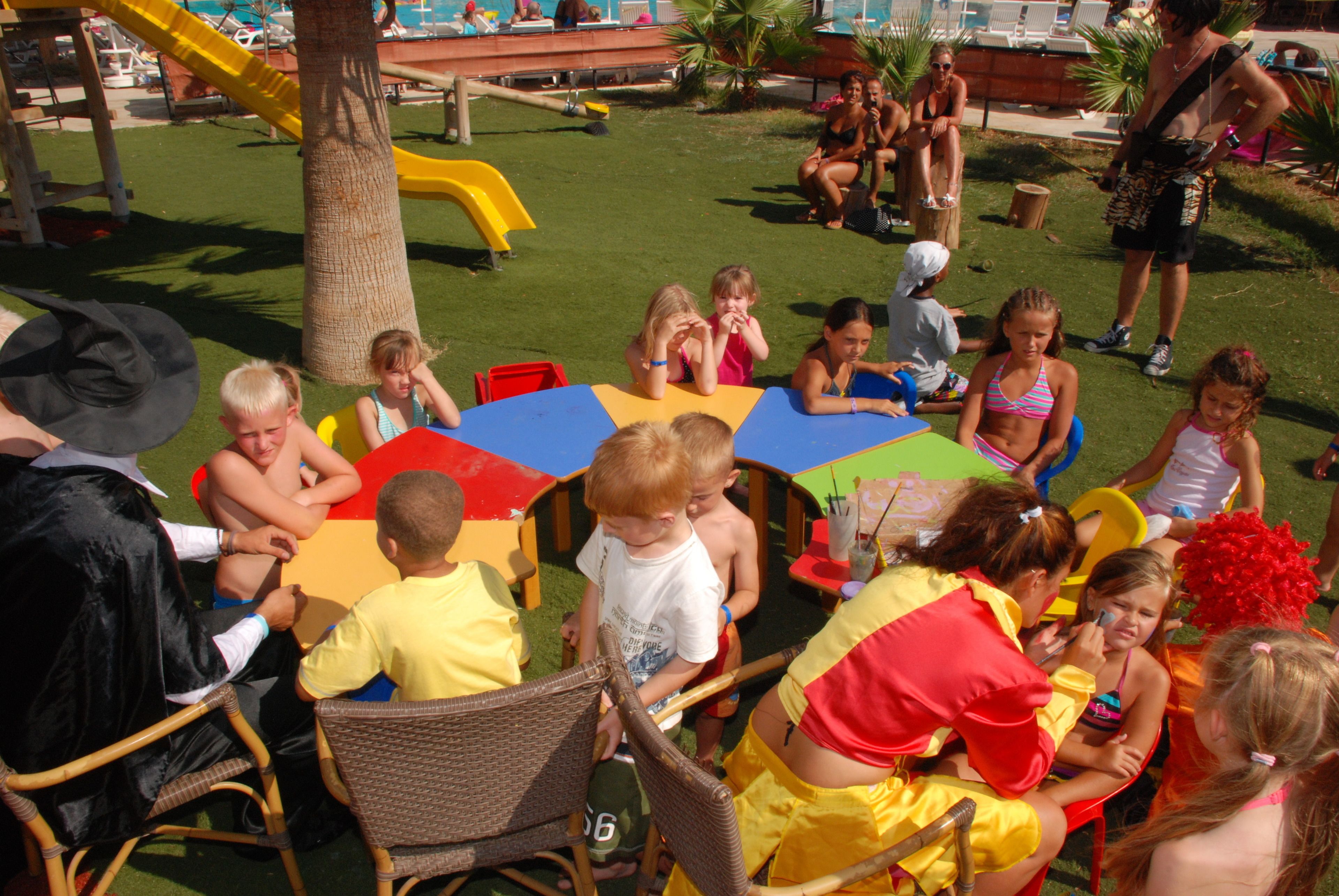 Niños y adultos sentados alrededor de una mesa colorida en un espacio al aire libre, con un parque infantil al fondo.