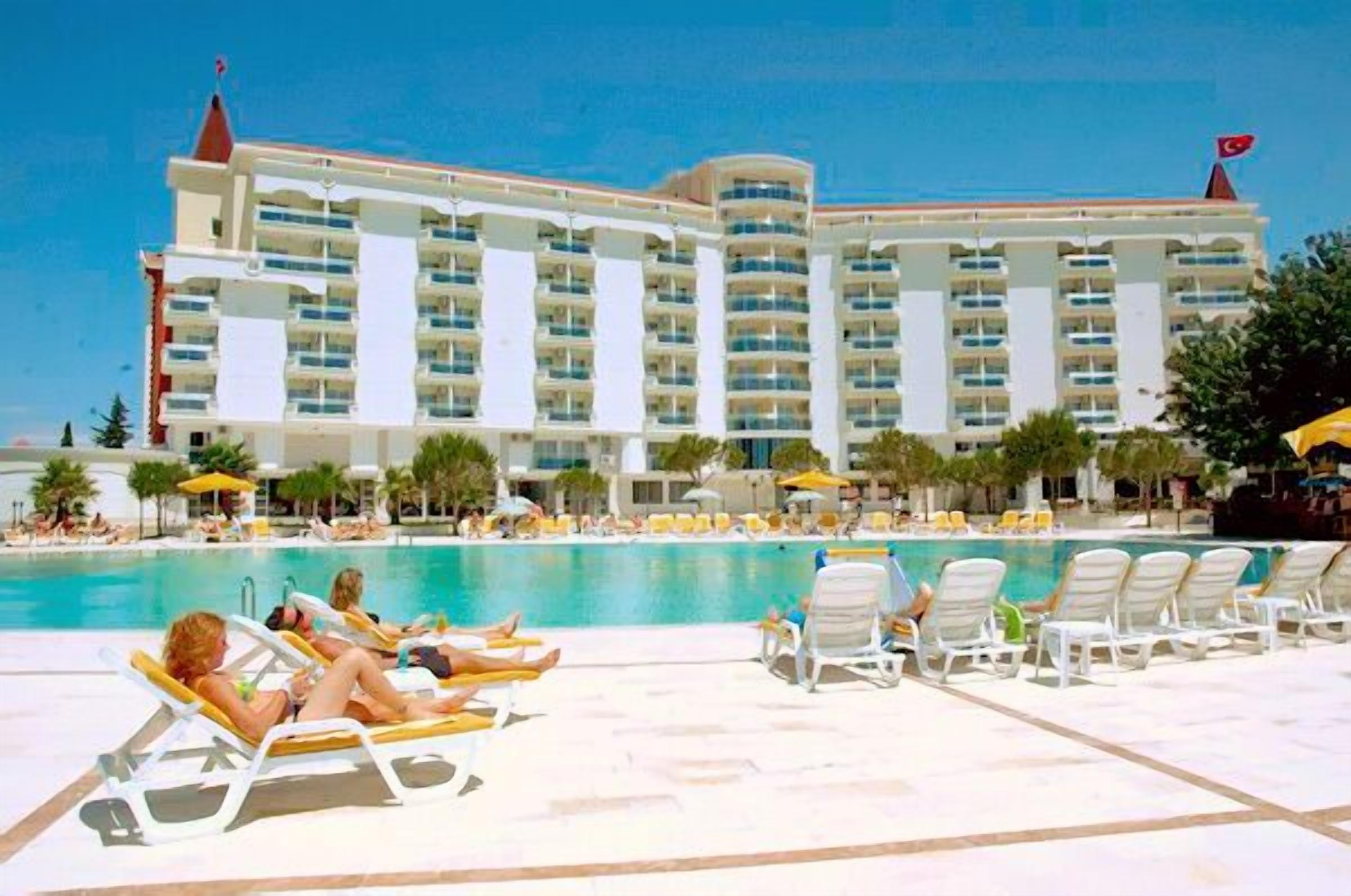 A hotel with multiple balconies overlooking a swimming pool, surrounded by sun loungers and umbrellas.