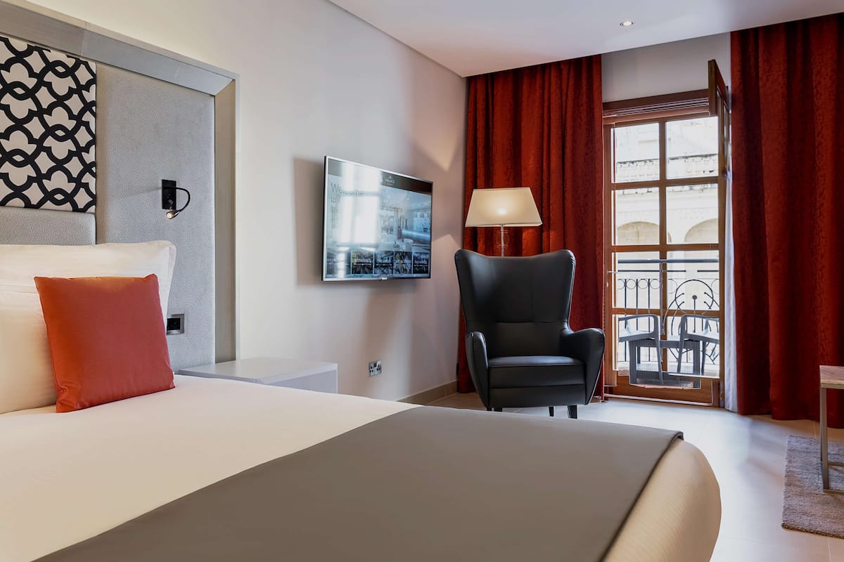 A hotel room with a large bed, a black armchair, a television mounted on the wall, and a window with red curtains.