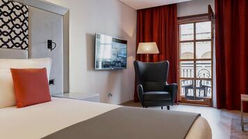 Et hotelværelse med en stor seng, en sort lænestol, et fjernsyn monteret på væggen og et vindue med røde gardiner.